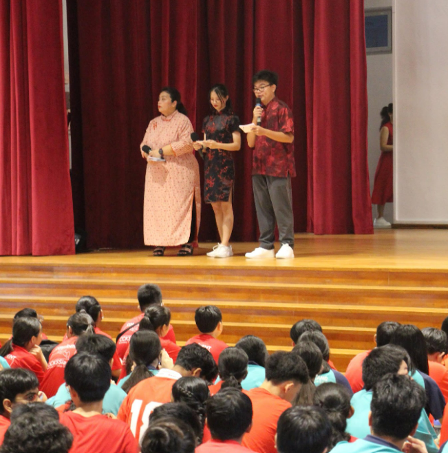 Three people stand on a stage with red curtains while a large crowd of students in teal and red shirts watches.